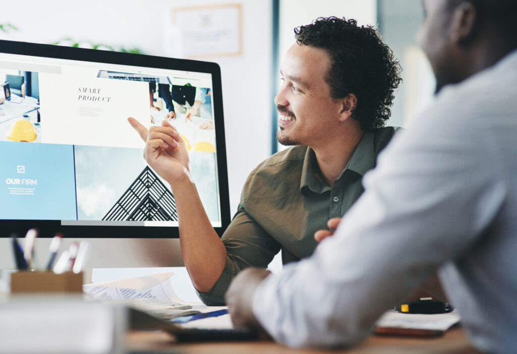 Two colleagues at a desk discussing a website mockup, with one man smiling and pointing at a desktop monitor displaying a webpage design.