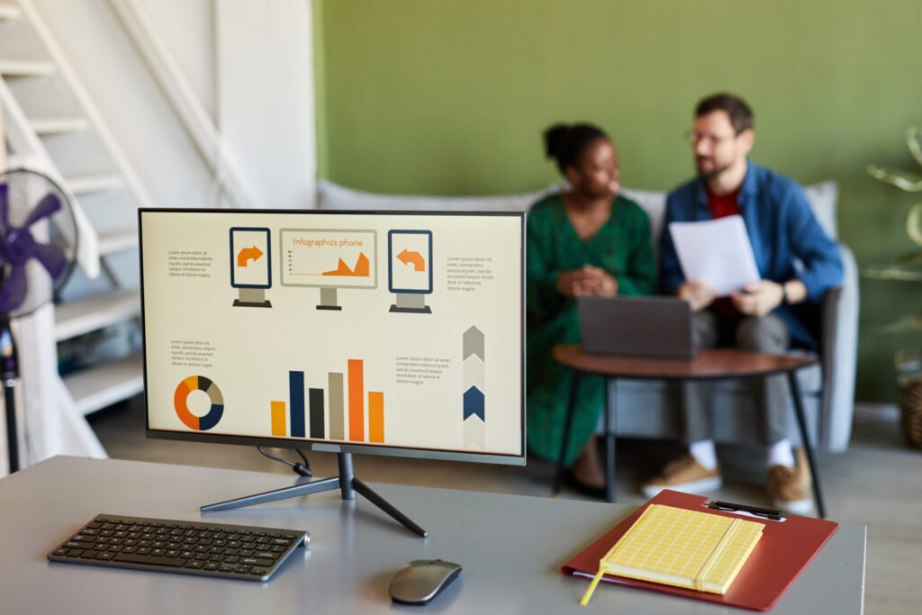 A computer monitor displaying business infographics, a pie chart, and a bar graph, with two colleagues reviewing documents in the blurred background.