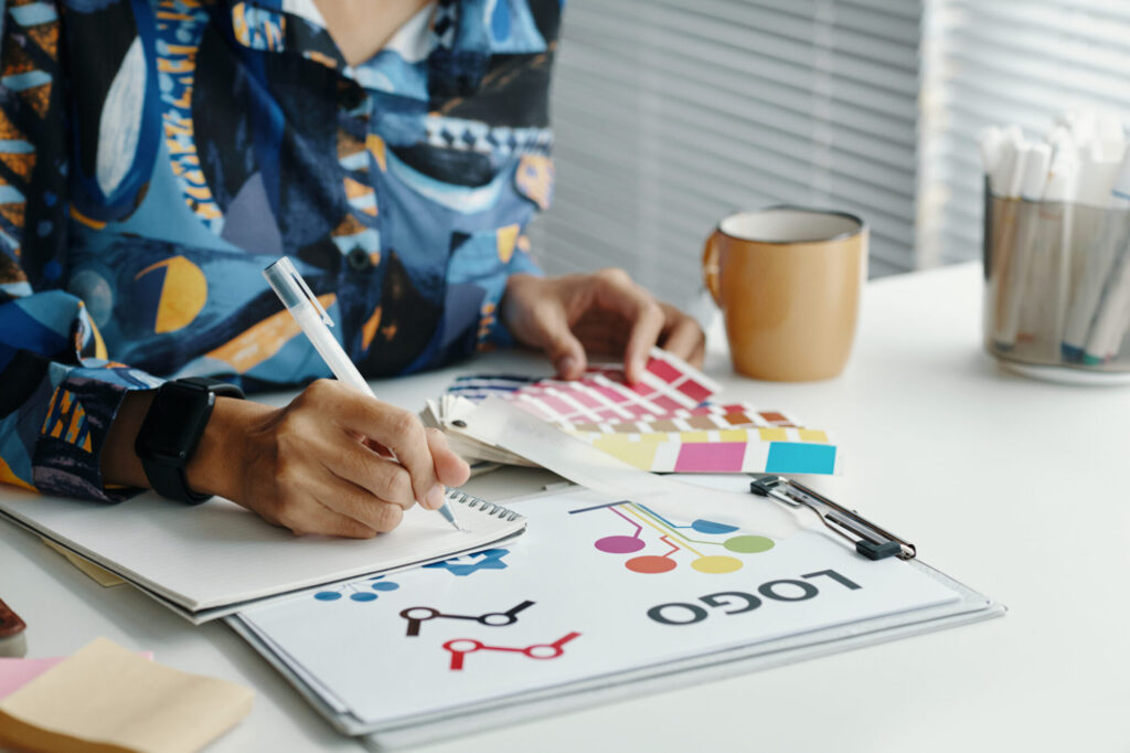 A graphic designer sitting at a desk sketching in a notebook, surrounded by color swatches and a printed logo design draft.