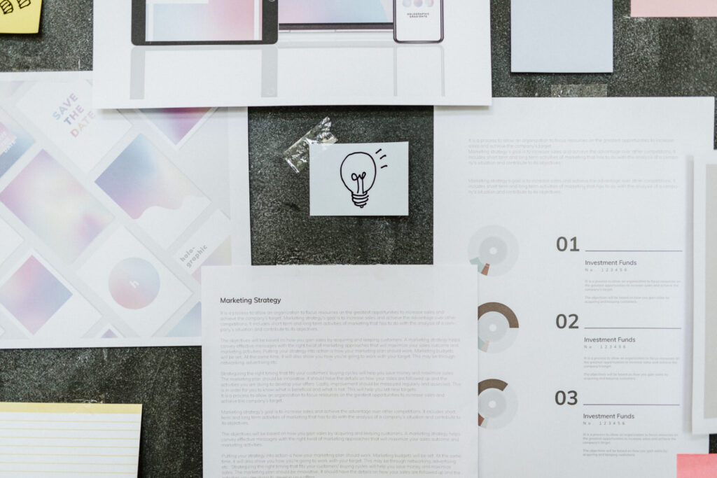 A flat lay of business planning documents, including a marketing strategy printout, design mockups, and a small sketch of a lightbulb.