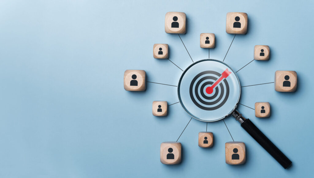 Wooden blocks with person icons connected by lines to a central magnifying glass that is focused on a red bullseye target.