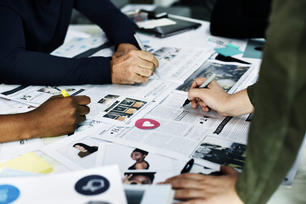 A close-up of multiple hands holding pens to review printed layout designs and articles scattered across a collaborative workspace.