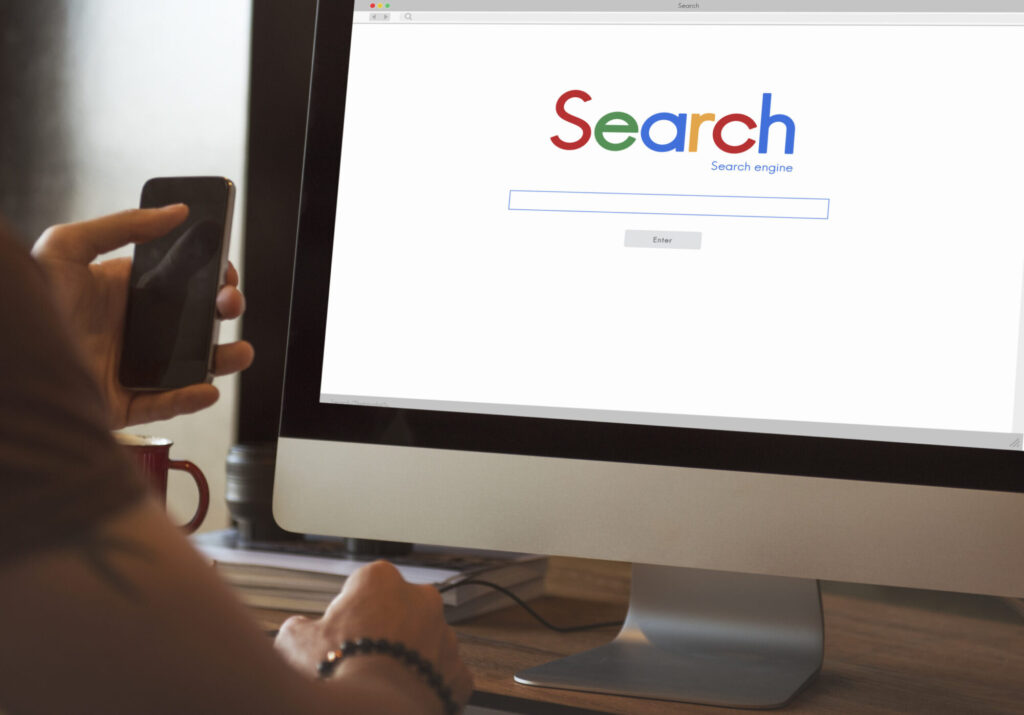 A person holding a smartphone while sitting at a desk, looking at a desktop computer monitor displaying a search engine homepage.
